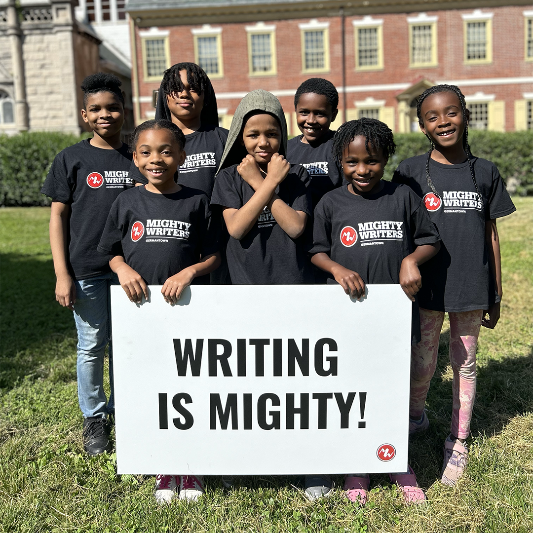 Seven children in black Mighty Writers t-shirts posing with a white "Writing is Mighty!" sign.
