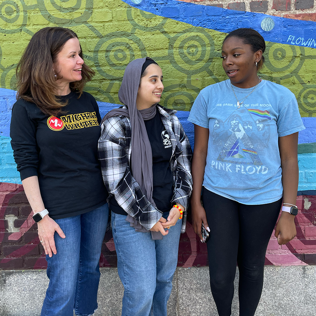 Two high school girls and one woman standing against a painted wall and looking and talking to each other.