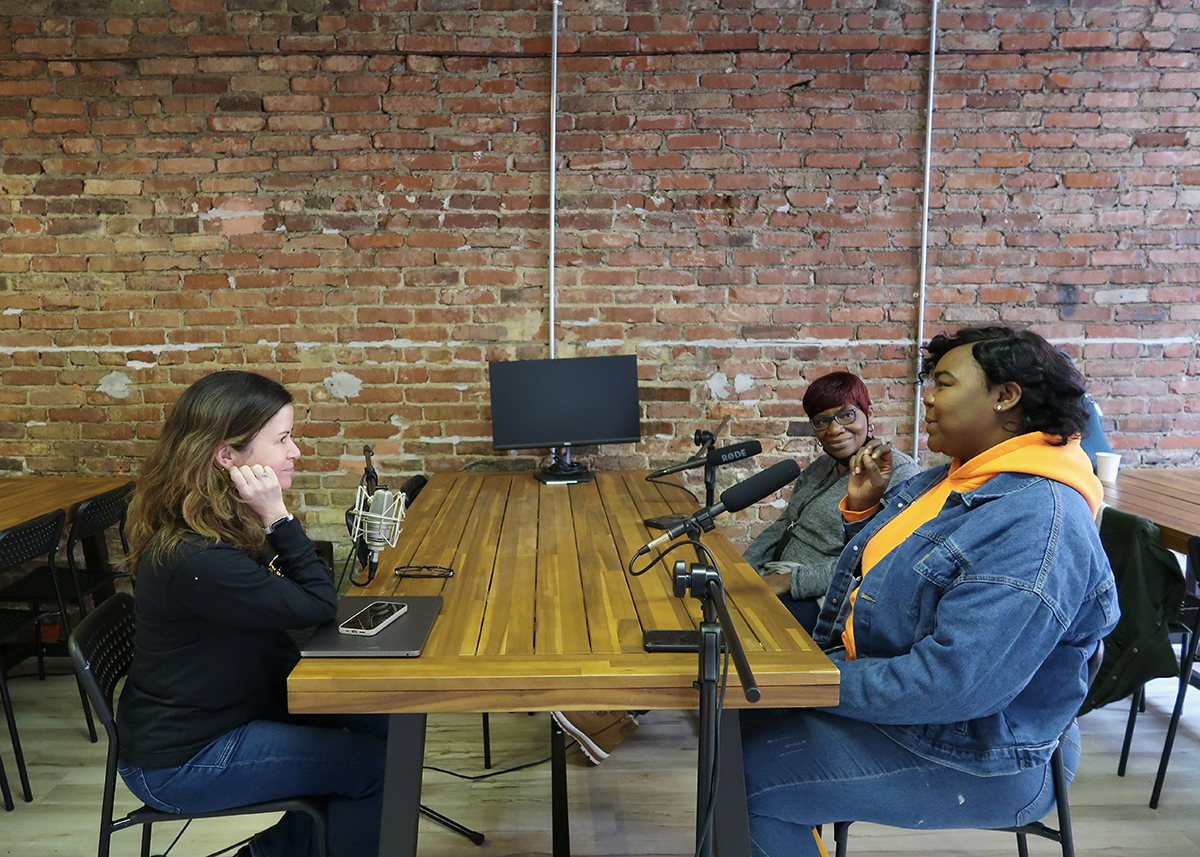 Three women seated at either side of a long table, two on one side with microphones in front of them and speaking, the other listening.