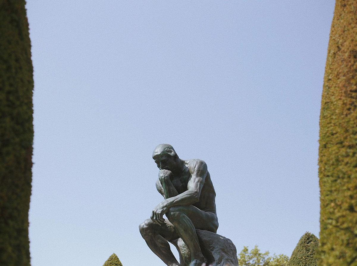 Rodin sculpture in the foreground with blue sky and trees in the background.