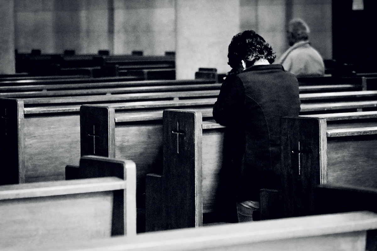 A black and white photo of someone kneeling in a church pew praying, and viewed from the back.